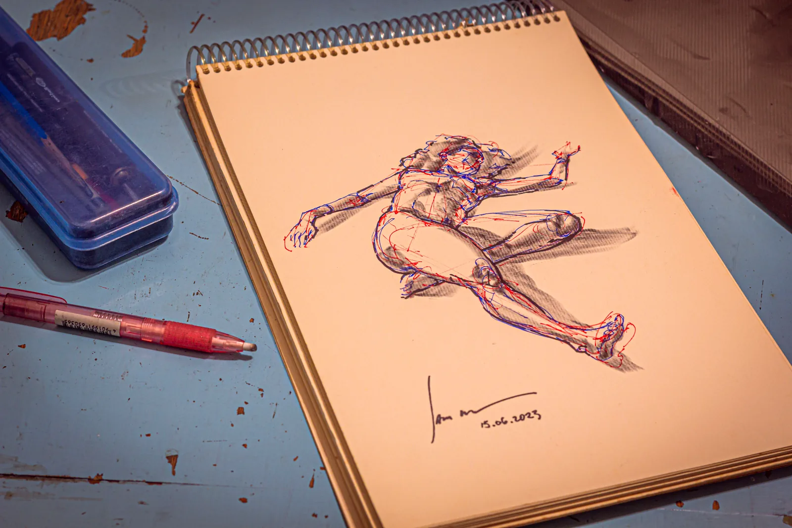 Caderno com desenho de figura humana e materiais de arte ao redor.